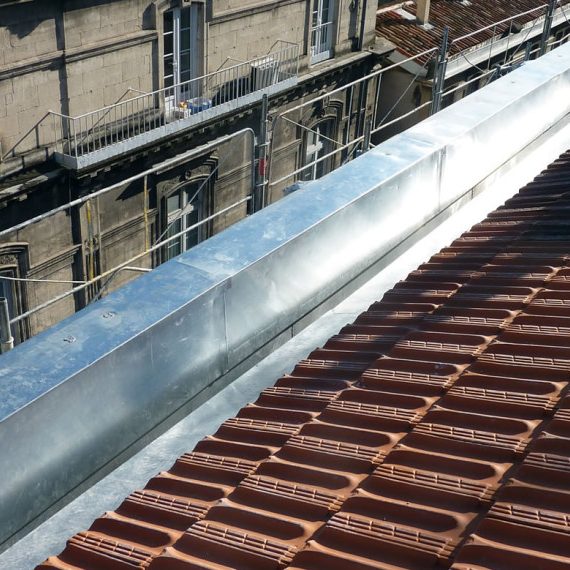 Toit en tuiles avec chéneaux zinc, vue d'un bâtiment en arrière-plan.