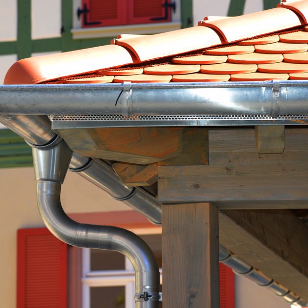 Nouvelle gouttière en métal sous un toit en tuiles, avec des volets rouges en arrière-plan.