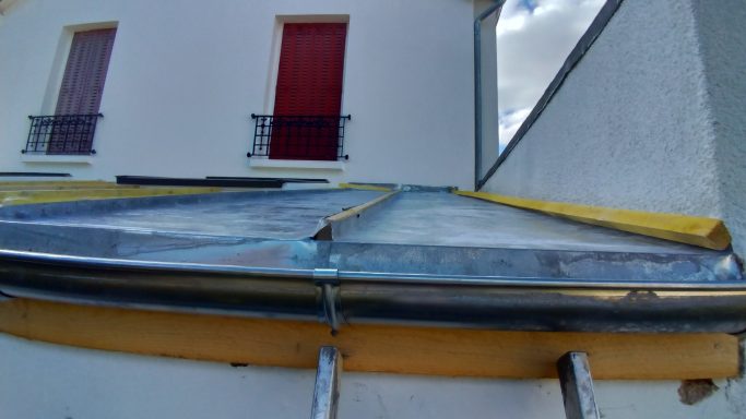 Toit en métal incliné avec bordure jaune, devant une maison à fenêtres rouges.