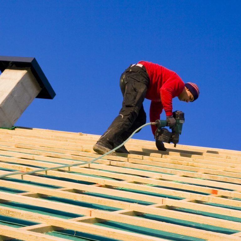 Ouvrier utilisant une perceuse sur un toit sous un ciel bleu.